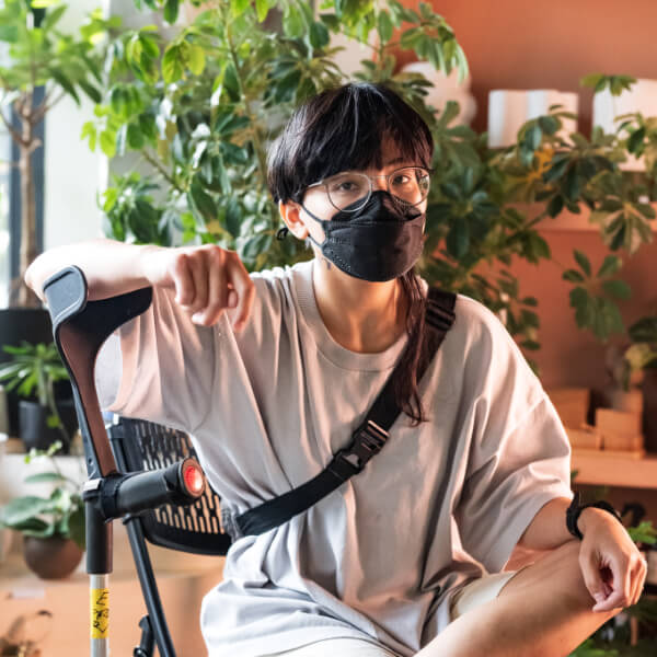A masked nonbinary Filipino person sits with his legs crossed, one arm casually leaning on his forearm crutch.
